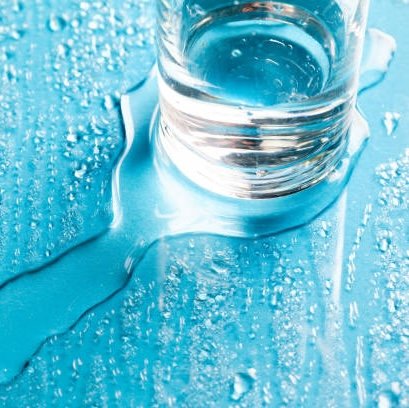 Glas mit Wasser auf blauer Oberfläche, umgeben von Wassertropfen, symbolisiert Feuchtigkeit und Pflege für trockene Haut, passend zum Thema Slugging in der Hautpflege.