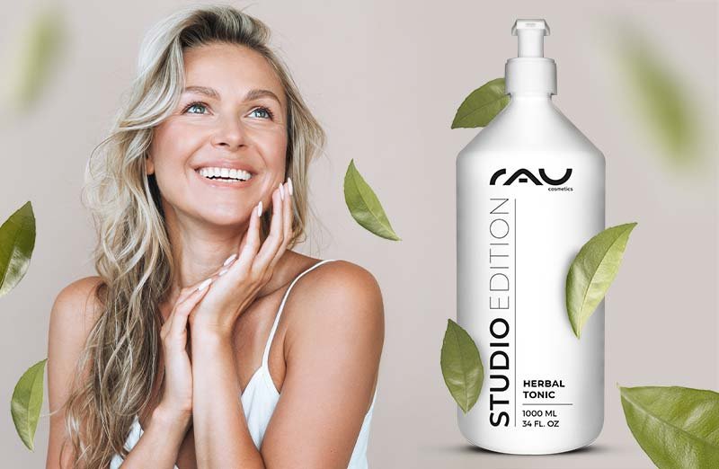 Lächelnde Frau mit langem Haar neben einer großen RAU Herbal Tonic-Flasche, umgeben von grünen Blättern.