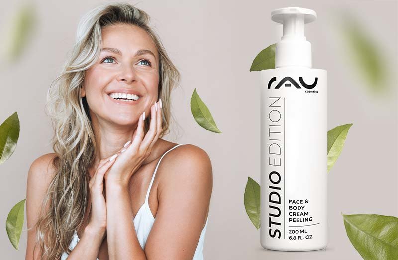 Lächelnde Frau mit welligem Haar und STUDIO EDITION Gesichts- & Körperpeelings mit 200 ML umgeben von grünen Blättern.