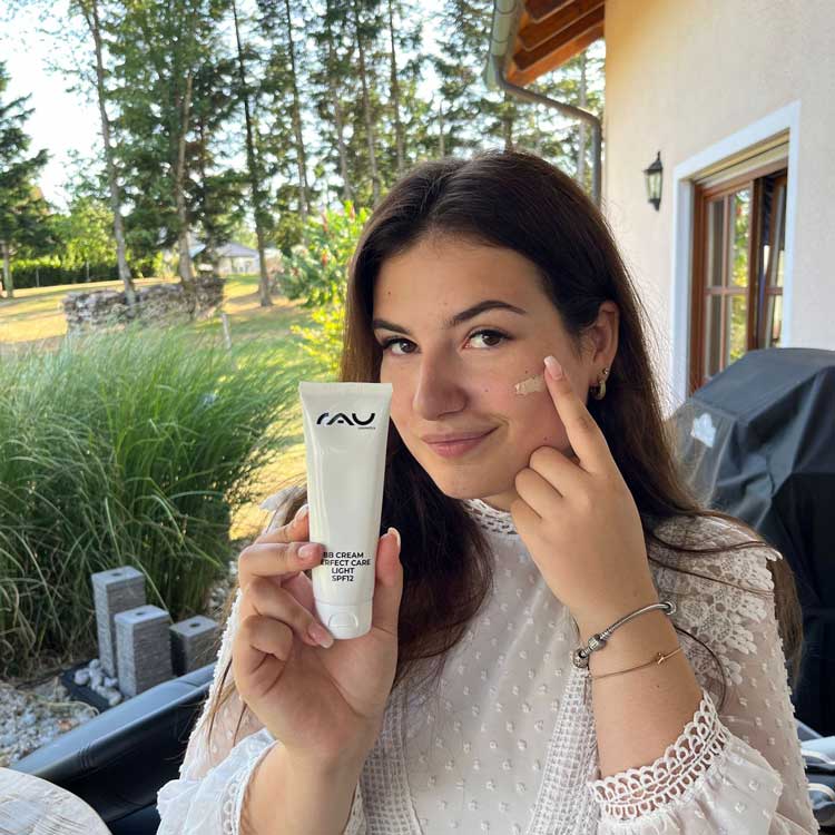 Junge Frau wendet RAU Hautpflegecreme in natürlicher Umgebung an, trägt eine stylische weiße Bluse.