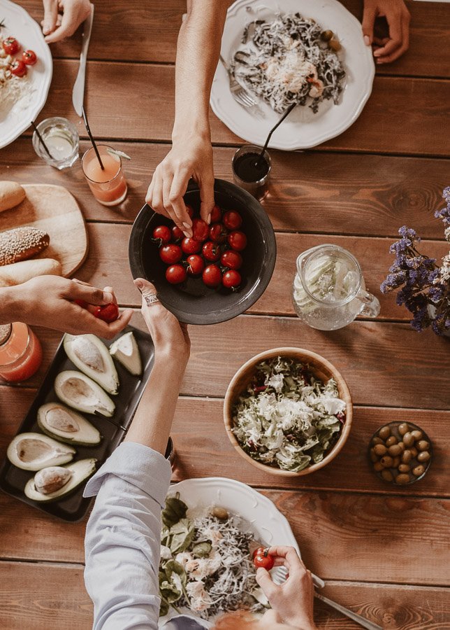 Hände reichen frische Tomaten und gesunde Speisen auf einem Holztisch, symbolisierend die Bedeutung von Ernährung für Anti-Aging und Hautgesundheit.