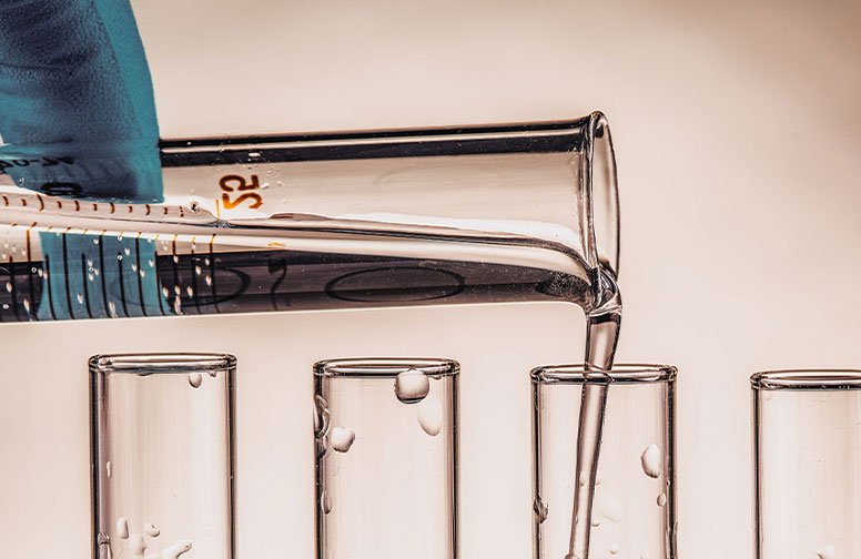 Glasbehälter mit Flüssigkeit wird in mehrere Reagenzgläser gegossen, symbolisiert die Herstellung von kolloidalem Silber für die Hautpflege.