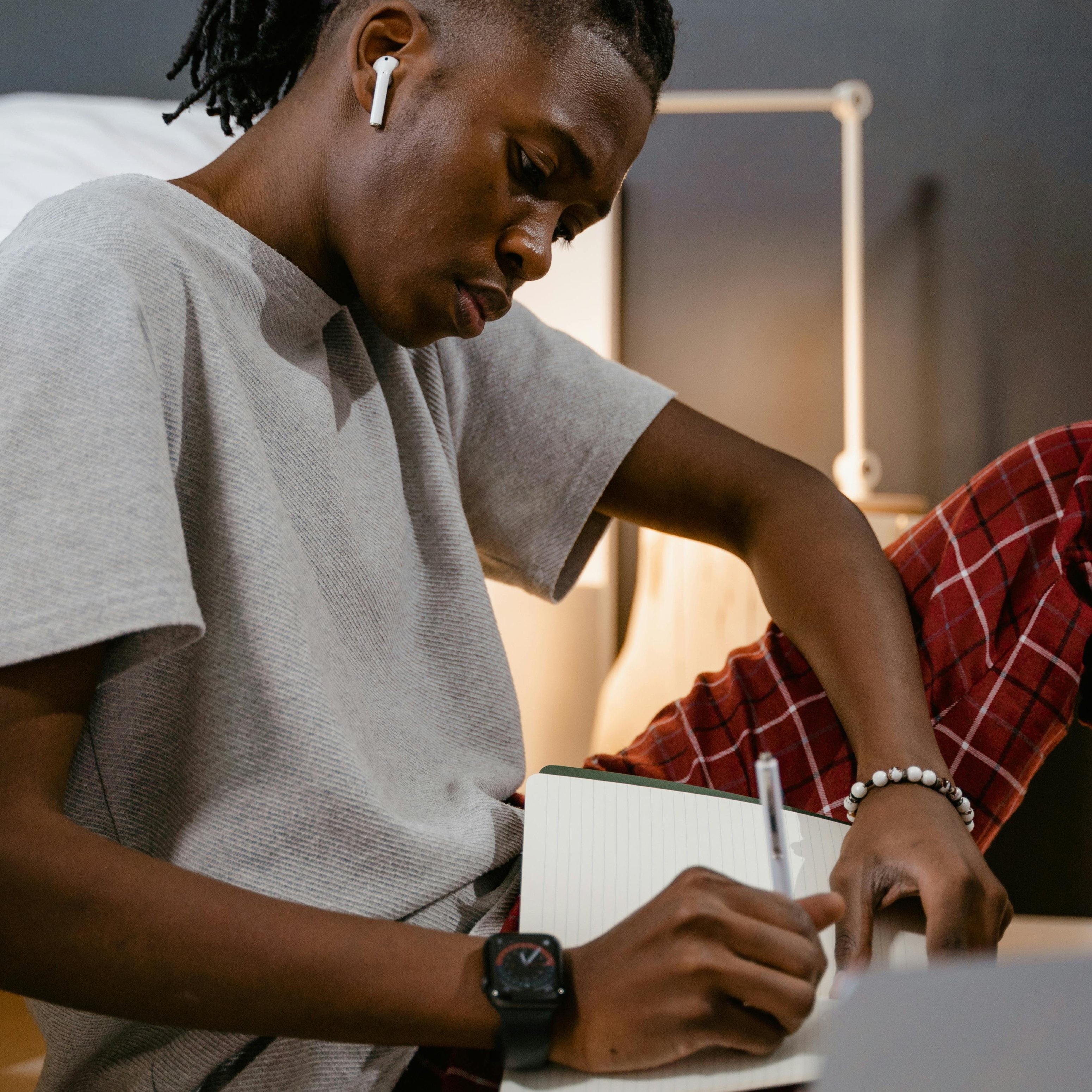 Junge Person mit AirPods, die in einem Notizbuch schreibt, in entspannter Umgebung mit warmem Licht, symbolisiert persönliche Reflexion und moderne Kommunikationsherausforderungen.