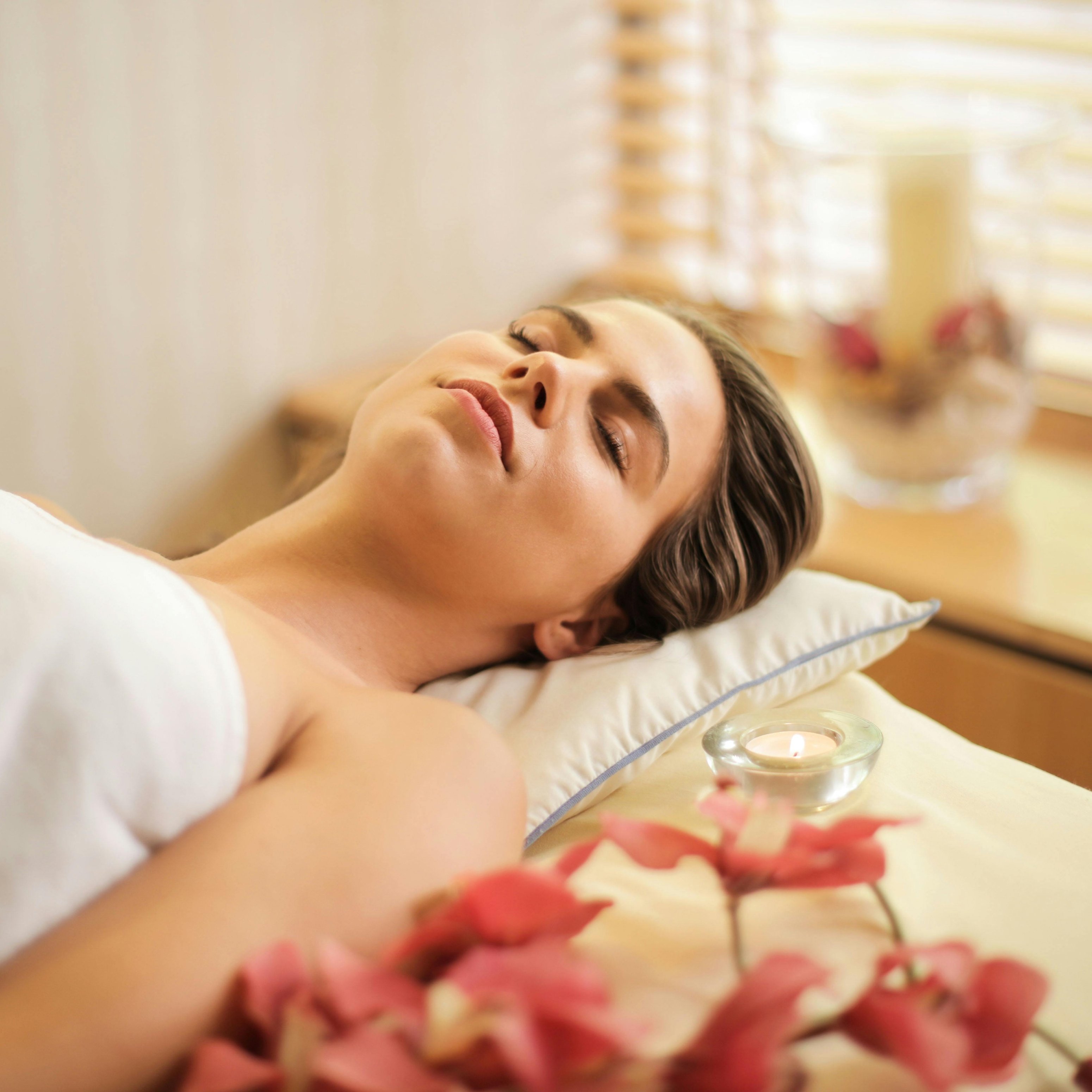 Frau entspannt während einer Gesichtsbehandlung mit Blumen und Kerze, repräsentiert die Vorteile der Buccal Massage für Hautverjüngung und Entspannung.