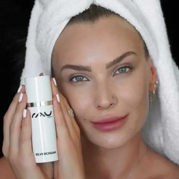 Eine Frau mit hellerer Haut lächelt, hält SILVER CREAM von RAU Cosmetics in der Hand, mit einem Handtuch auf dem Kopf.